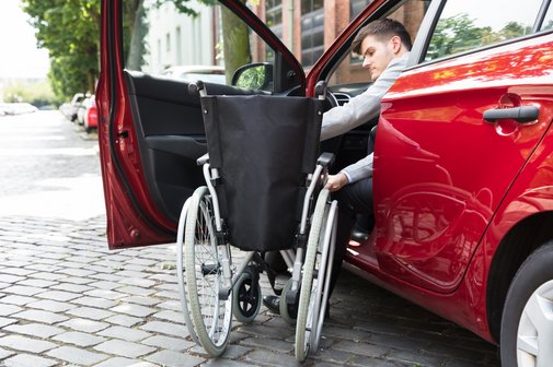 Autofahrer mit Behinderung Ein junger Mann steigt von seinem Rollstuhl in sein Auto ein und setzt sich auf den Fahrersitz.