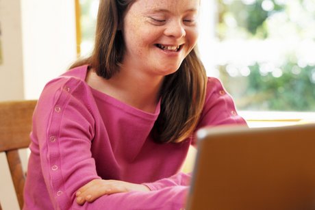 Eine junge Frau mit Downsyndrom sitzt an einem Tisch, vor sich ein aufgeklapptes Notebook.