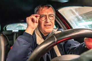 Ein älterer Mann am Steuer eines Autos. Er fasst sich an die Brille, sieht konzentriert oder überrascht aus. 