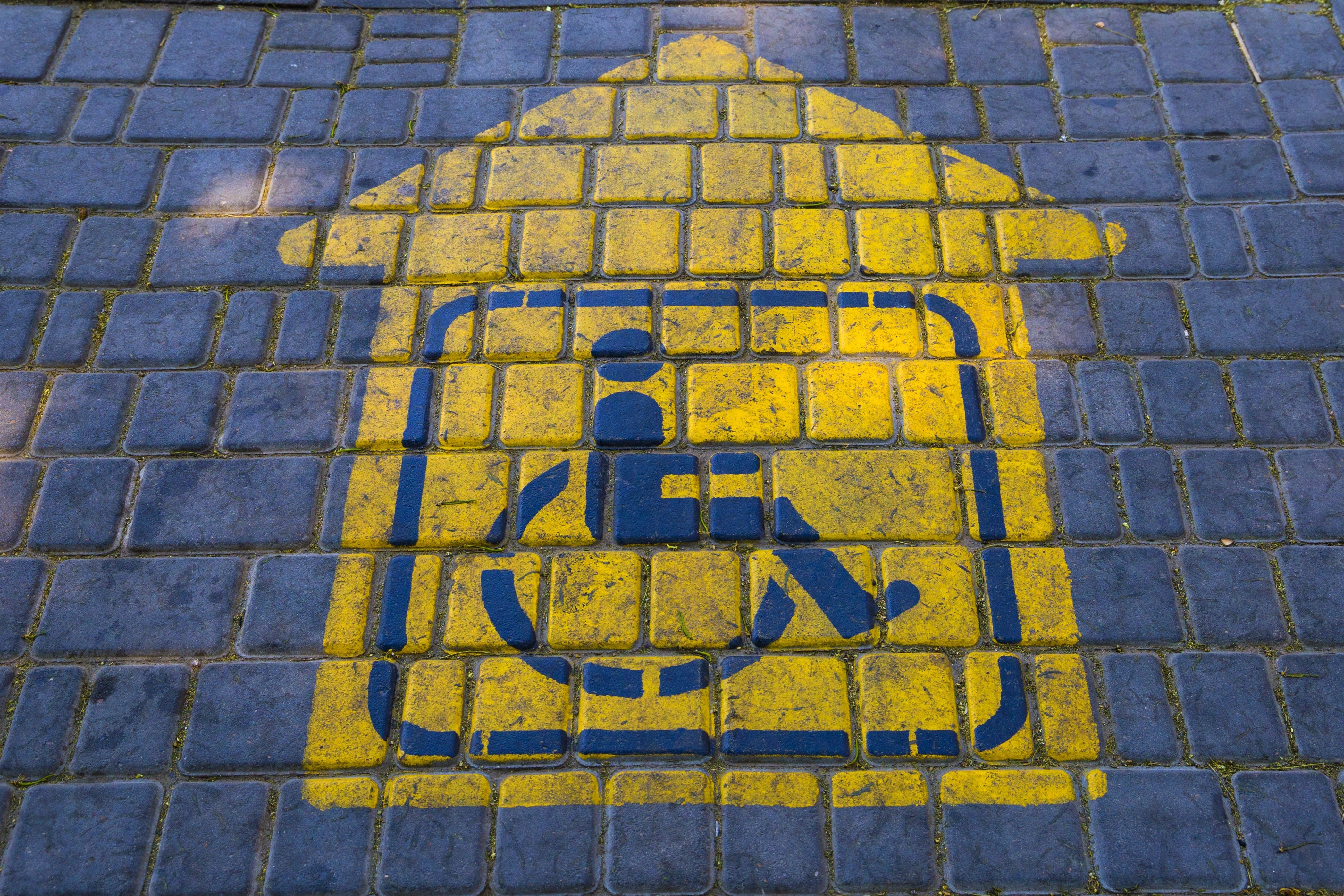 Ein mit gelber Farbe auf den Boden aufgemaltes Piktogramm auf einem gepflasterten Platz zeigt mit einem Pfeil an, wo sich die Parkplätze für Rollstuhlfahrer und Menschen mit Behinderung befinden. 