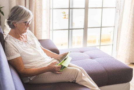 Eine ältere Frau sitzt auf einem Sofa, liest in einem Buch.