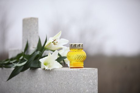 Ein Grabstein, darauf liegen weiße Lilien und ein Grablicht in einem gelben Kerzenhalter