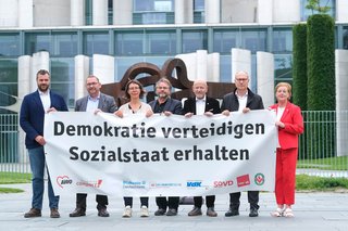 Mitglieder des Bündnisses vor dem Bundeskanzleramt; sie halten ein großes Transparent in den Händen mit der Aufschrift "Demokratie verteidigen - Sozialstaat erhalten"