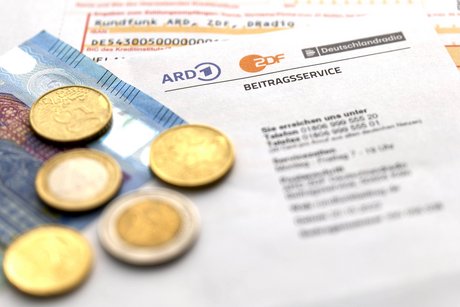 Ein Schreiben des Rundfunkgebühren-Beitragsservice, darauf liegen Münzen und ein 20-Euro-Schein, darunter ein Überweisungsträger