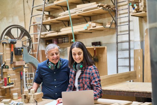 Ältere auf dem Arbeitsmarkt Ein älterer Mann und eine Frau in einer Schreinerei, sie beugen sich gemeinsam über ein Notebook, im Hintergrund Holz, Werkzeuge und Maschinen
