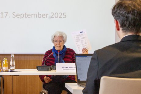 SoPoK September 2025 Ulrike Mascher sitzt an einem Tisch und hält ein Dokument in die Höhe. Vor ihr steht ein Namensschild, hinter ihr wird etwas an eine Leinwand gebeamt.
