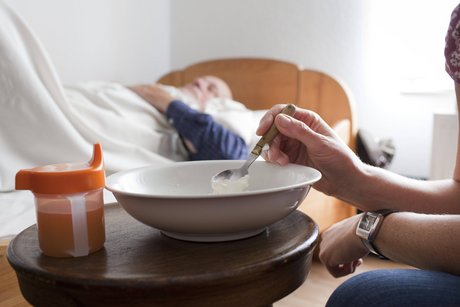 Ein älterer Mann liegt in einem Pflegebett. Im Vordergrund sieht man eine Frau sitzen, die einen Löffel in einen Teller mit Suppe taucht, um ihn zu füttern. Auf dem Tisch steht auch eine Schnabeltasse. 