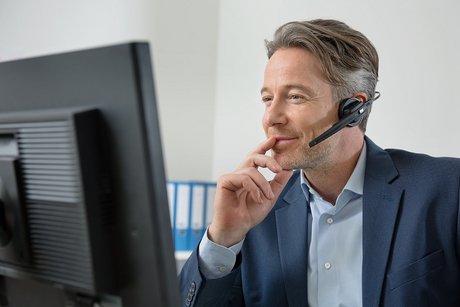 Mann mit Headset vor einem Computerbildschirm, er telefoniert, führt nachdenklich die Finger zum Mund. 