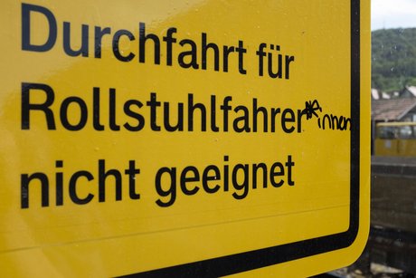 Ein Gelbes Hinweisschild mit dem Text "Durchfahrt für Rollstuhlfahrer nicht geeignet". Mit einem Edding hat jemand das Wort Rollstuhlfahrer ergänzt zu Rollstuhlfahrer*innen.