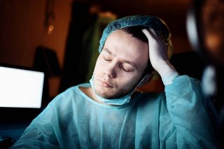 Ein müde wirkender Mann in OP-Kleidung sitzt mit geschlossenen Augen, er greift sich an den Kopf.