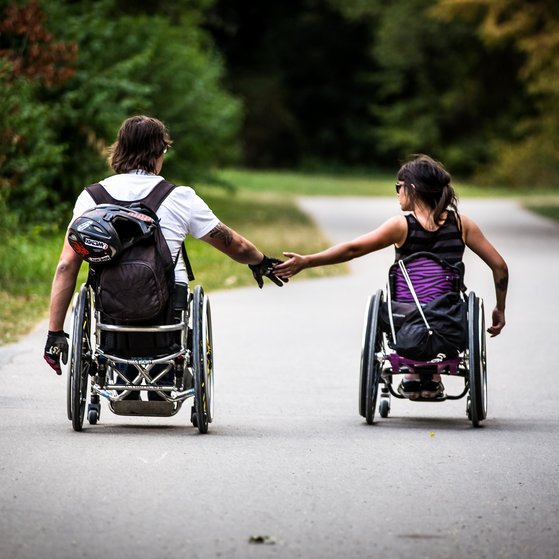 Ein junger Mann und eine junge Frau im Rollstuhl, sie fahren nebeneinander, halten sich an der Hand. Das Foto zeigt Lisa Schmidt und David Lebuser; beide sind Wheelchairskater und testen den Skatepark in München.