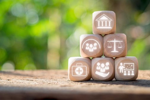 Soziale Sicherheit Sechs Würfel mit unterschiedlichen Symbolen sind aufeinander gestapelt, unter anderen mit Icons für Gesundheit, Recht, Gemeinschaft und Bildung.