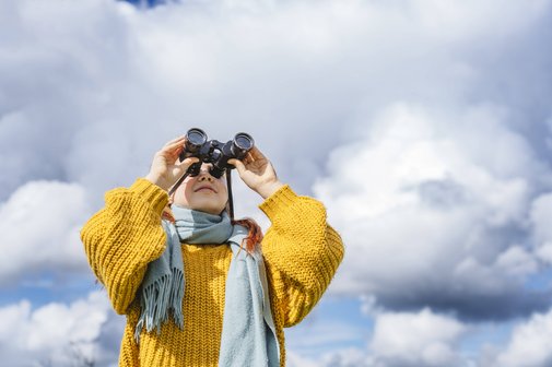 Eine Frau in einer gelben Strickjacke vor blauem Himmel mit weißen Wolken, sie blickt durch ein Fernglas.