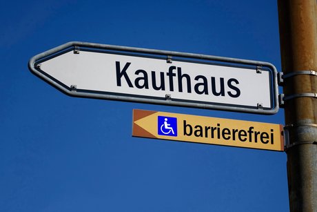 Das Bild zeigt einen Wegweiser mit der Aufschrift "Kaufhaus", darunter ein kleineres Hinweisschild mit der Aufschrift "barrierefrei" und einem Rollstuhl-Piktogramm