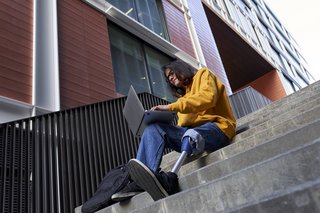 Ein junger Mann mit einer Bein-Prothese sitzt auf einer Treppe, arbeitet mit seinem Laptop.