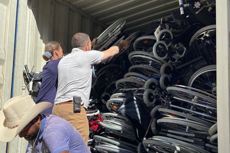 Zwei Männer und eine Frau beladen einen Schiffscontainer mit ausrangierten Rollstühlen, der gesamte Container ist vollgestapelt mit den Rollstühlen. 