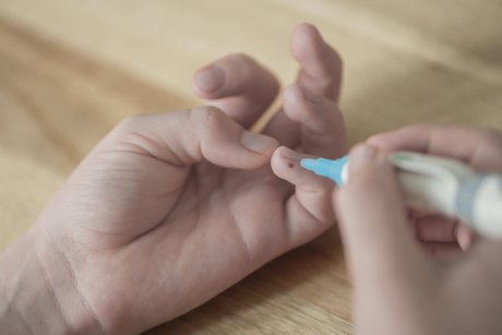 Das Bild zeigt eine Kinderhand, deren kleiner Finger einen Tropfen Blut zeigt. In der rechten Hand hält das Kind die Lanzette als Stechhilfe, hat sich gerade gepiekst, um den Blutzucker zu bestimmen.