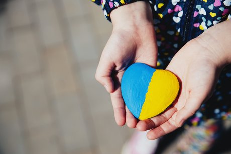 Hand eines Kindes hält einen herzförmigen Stein, der zur Hälfte blau und zur Hälfte gelb angemalt ist in den Farben der ukrainischen Flagge