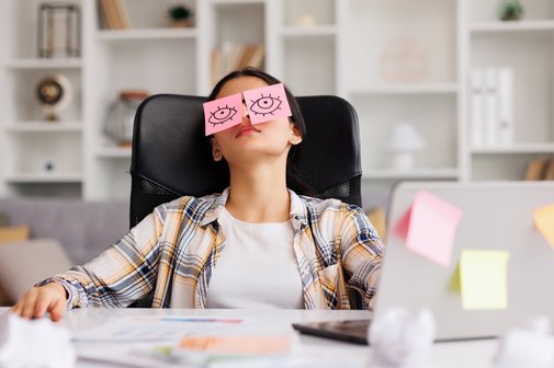 Belastung im Arbeitsleben Eine junge Frau sitzt am Schreibtisch in einem Büro, sie hat den Kopf zurückgelegt. Auf ihren geschlossenen Augen liegen Post-it-Zettel mit aufgemalten offenen Augen.