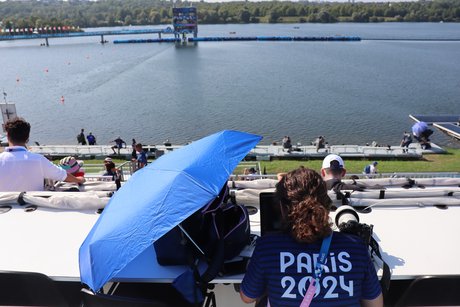 Menschen sitzen auf einer Tribübe mit Blick das Wassersportstadion Vaires-sur-Marne . Eine Frau trägt ein T-Shirt mit dem Rückenaufdruck "Paris 2024"