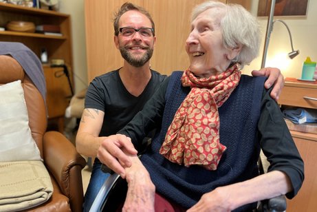 Besuch im Seniorenheim Dirk Angele und Anita Göbels, die Bewohnerin des Seniorenheims, in dem Dirk Angele ehrenamtlich tätig ist. Sie sitzt im Rollstuhl, er sitzt hinter ihr, legt eine Hand auf ihre Schulter und die andere auf ihren Arm.