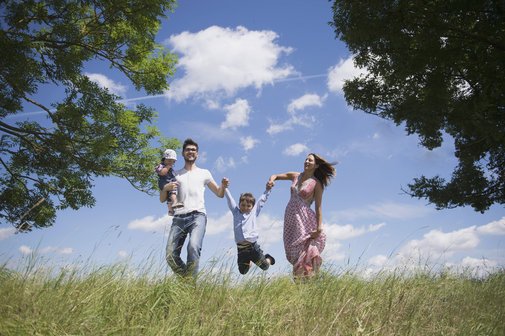 Familie Eine junge Familie mit zwei kleineren Kindern auf einer Wiese vor blauem Himmel, der Vater trägt ein Kind auf dem Arm, das andere Kind halten die Eltern zwischen sich und lassen es "fliegen".