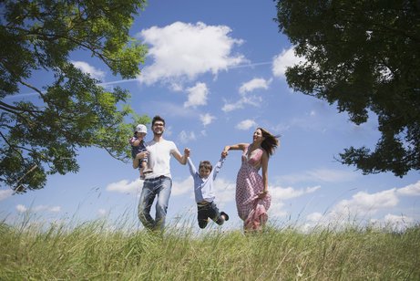 Familie Eine junge Familie mit zwei kleineren Kindern auf einer Wiese vor blauem Himmel, der Vater trägt ein Kind auf dem Arm, das andere Kind halten die Eltern zwischen sich und lassen es "fliegen".
