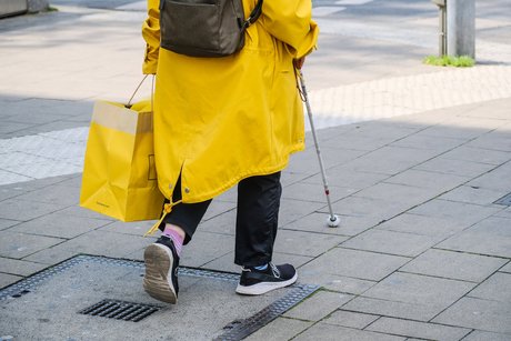 Person in gelben Regenmantel läuft mit einem Blindenstock entlang eines Blindenleitsystems 