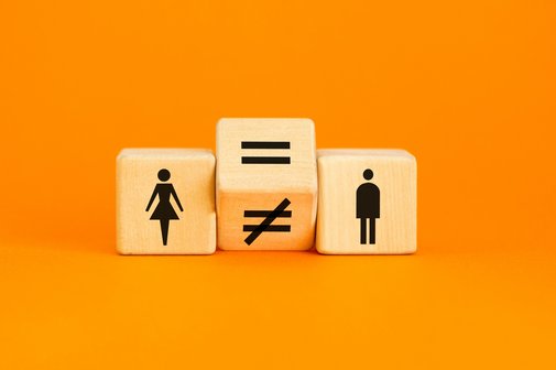 Entgeltgleichheit Drei Würfel liegen nebeneinander vor orangenem Hintergrund. Einer zeigt ein Piktogramm einer Frau, einer das eines Mannes. Dazwischen liegt ein gekippter Würfel auf der Kante, der ein Gleichheits- und ein Ungleichheitszeichen anzeigt.