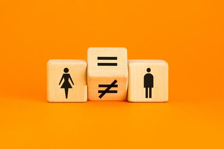 Entgeltgleichheit Drei Würfel liegen nebeneinander vor orangenem Hintergrund. Einer zeigt ein Piktogramm einer Frau, einer das eines Mannes. Dazwischen liegt ein gekippter Würfel auf der Kante, der ein Gleichheits- und ein Ungleichheitszeichen anzeigt.