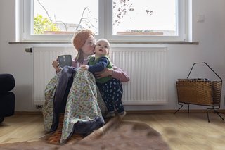 Eine Mutter und ihr Kleinkind sitzen an einem Heizkörper, sie tragen warme Kleidung, die Mutter hält eine Teetasse in der Hand. Sie gibt dem Kind einen Kuss auf die Wange. 