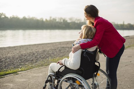 Eine ältere Frau im Rollstuhl am Ufer eines Flusses; eine jüngere Frau steht hinter ihr, legt liebevoll den Arm um sie.