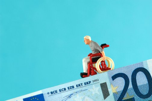 Pflege und Armut Symbolbild: Ein 20-Euro-Schein bildet eine abschüssige Rampe, auf der eine Mini-Figur eines alten Mannes im Rollstuhl bergab rollt