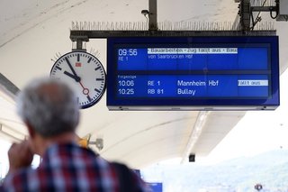 Rückenansicht eines älteren Mannes an einem Bahnstein am Bahnhof Koblenz, man sieht die digitale Anzeigetafel mit den angekündigten Zügen sowie eine Bahnhofsuhr.