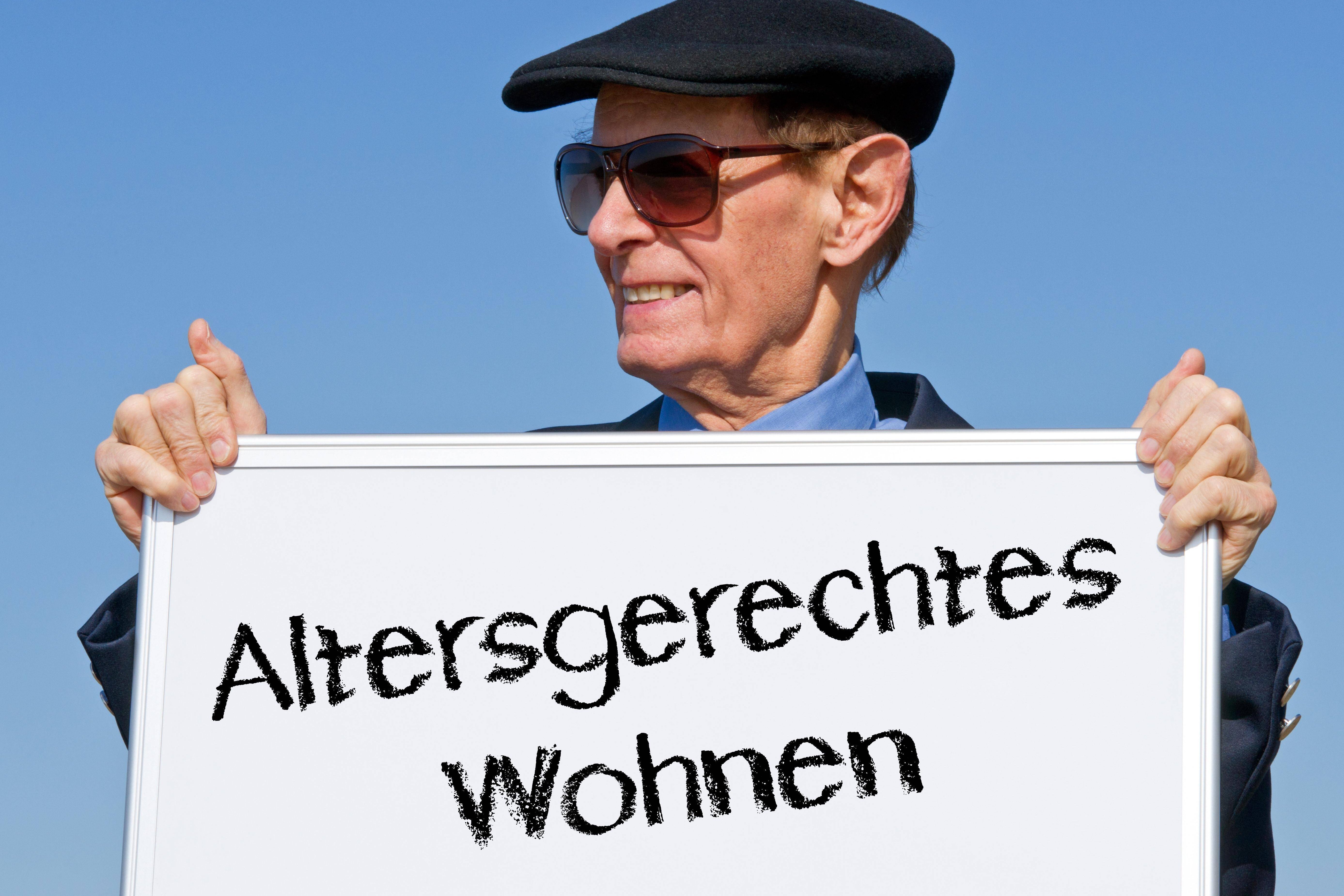 Senior mit Schirmmütze und Sonnenbrille hält ein Schild in beiden Händen, auf dem steht "Altersgerechtes Wohnen"