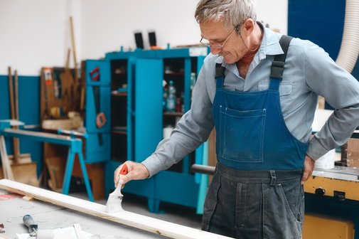 Arbeitsmarkt für Ältere Ein älterer Handwerker mit grauem Haar und Brille streicht eine Leiste mit weißer Farbe, hat eine Hand in den Rücken gestützt, trägt einen Blaumann