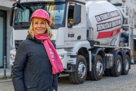 VdK-Präsidentin Verena Bentele steht vor dem großen Betonmischer, sie trägt einen rosa Schal mit Aufdruck "Gegen soziale Kälte" und einen rosa Bauarbeiterinnen-Helm