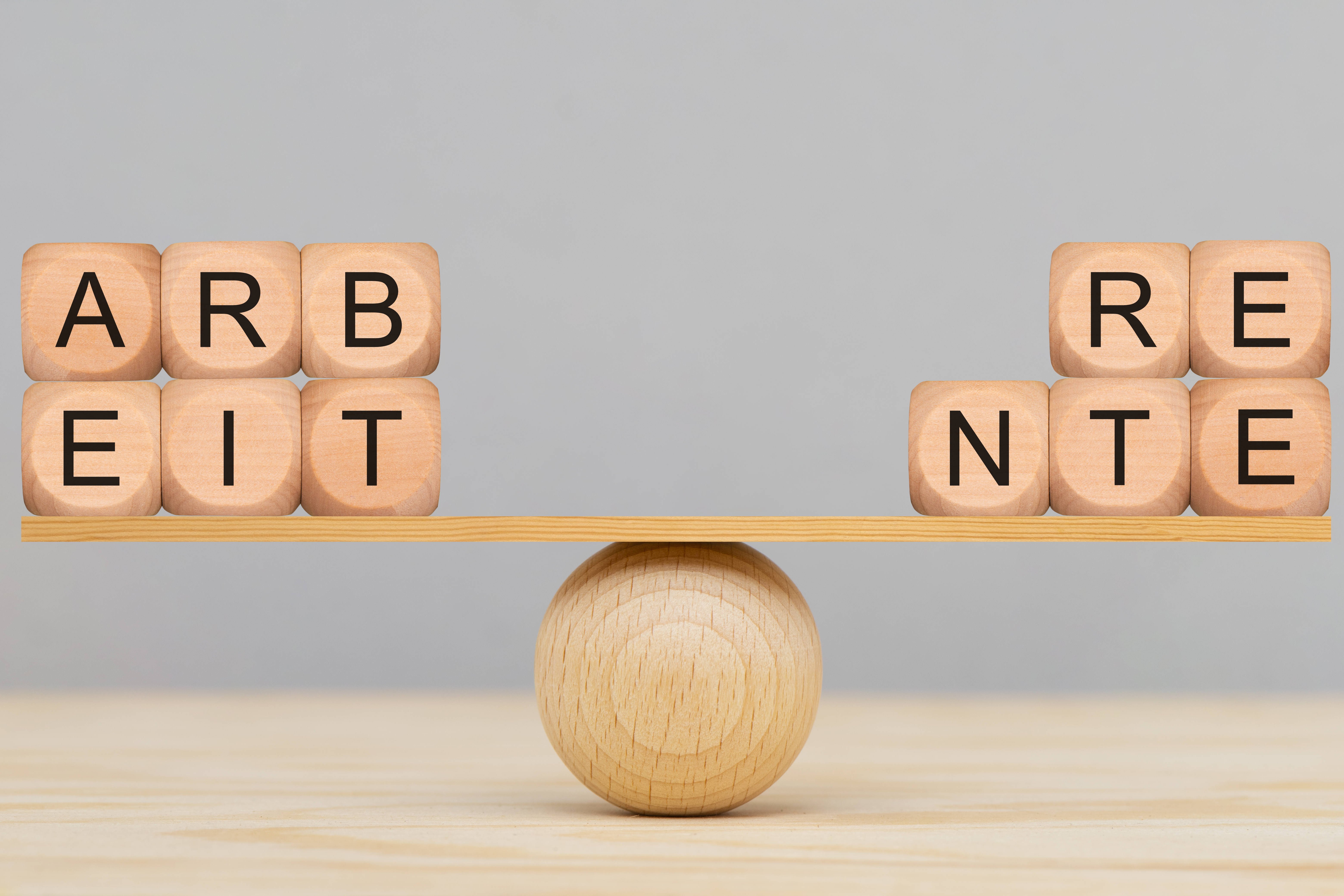 Holzbuchstaben werden auf einer Wippe aus einer Holzkugel und einem kleinen Holzbrett ausbalanciert. Auf der linken Seite bilden die Buchstabenwürfel das Wort "Arbeit", auf der rechten Seite "Rente".