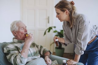 Eine junge Frau mit steht neben einem Senior, der in einem Sessel sitzt und Saft aus einem Glas trinkt. Sie hält ein Medikamentenröhrchen in der Hand. 