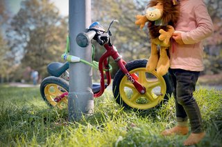 Ein Laufrad für Kleinkinder ist an einem Laternenmast mit einem Fahrradschloss festgemacht. Im Vordergrund sieht man ein Kind, das einen Plüsch-Löwen im Arm hält.