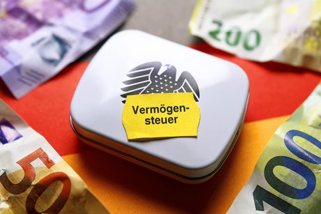 Die Bildmontage zeigt eine Box mit einem Bundesadler darauf. Auf ihr klebt ein gelbes Preisschild mit der Aufschrift "Vermögensteuer". Darum herum liegen Euro-Geldscheine.