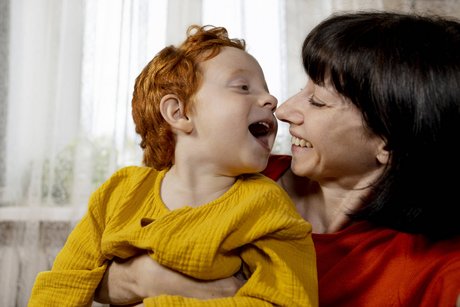 Mutter und Kleinkind, die Mutter hält das Kind fest, sie lachen beide fröhlich, sind sehr nah und zugewandt. 