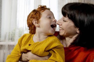 Mutter und Kleinkind, die Mutter hält das Kind fest, sie lachen beide fröhlich, sind sehr nah und zugewandt. 