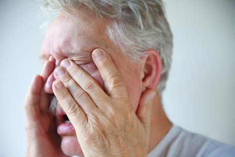 Ein Mann fasst sich mit beiden Händen ins Gesicht und an die Nase, er guckt leidend, hat die Augen geschlossen. 