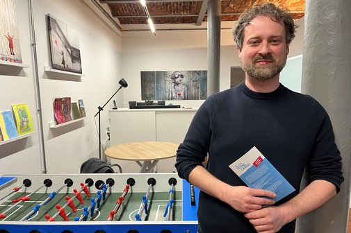 Benjamin Salzmann Benjamin Salzmann im Young Carer Zentrum in Berlin-Moabit, er steht an eine Säule gelehnt und hält einen Flyer in den Händen, hinter ihm steht ein Kickertisch.