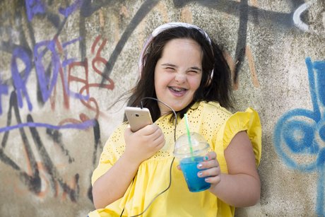 Junge Frau mit Downsyndrom, sie lacht in die Kamera, hält ihr Smartphone in der einen Hand und einen Plastikbecher mit einem Getränk in der anderen. Sie trägt Kopfhörer.