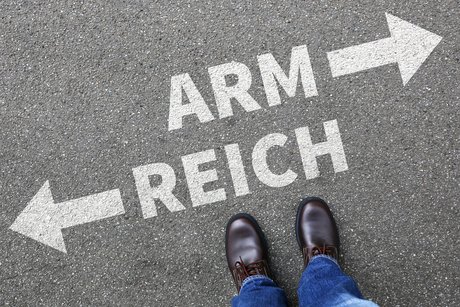 Füße auf Asphalt, darauf steht in weißer Schrift aufgesprüht "Arm" mit einem Pfeil nach rechts und "Reich" mit einem Pfeil nach links