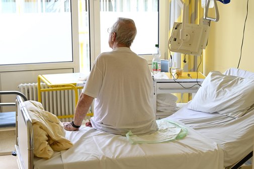 Senior im Krankenhaus Symbolbild: Ein älterer Patient sitzt in seinem Krankenhauszimmer auf seinem Bett, man sieht ihn in der Rückenansicht