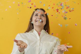 Eine Frau in weißer Bluse fängt lachend buntes Konfetti auf, sie steht vor einem gelben Hintergrund.