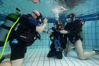 Janis McDavid beim Tauchen im Schwimmbecken. Zwei Mitglieder des Tauchclubs geben Hilfestellung. 